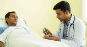 একজন বাংলাদেশী রোগী বেডে শুয়ে আছেন, একজন বাংলাদেশী ডাক্তার রোগীর হাত থেকে রক্তের শরর্করা পরীক্ষা করছেন। আকটি আয়তকার ছবি